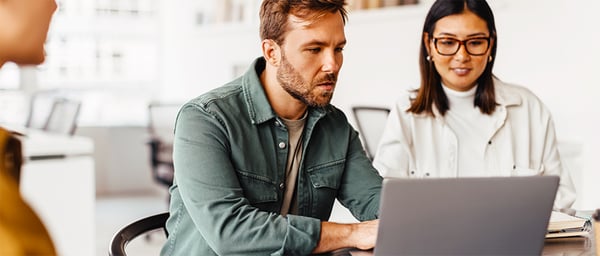 Two professionals work together at a laptop in a bright modern office. A man in a green shirt focuses on the screen while a woman wearing glasses looks on attentively. Their collaborative posture and shared workspace suggest teamwork, problem-solving, and productive discussion.